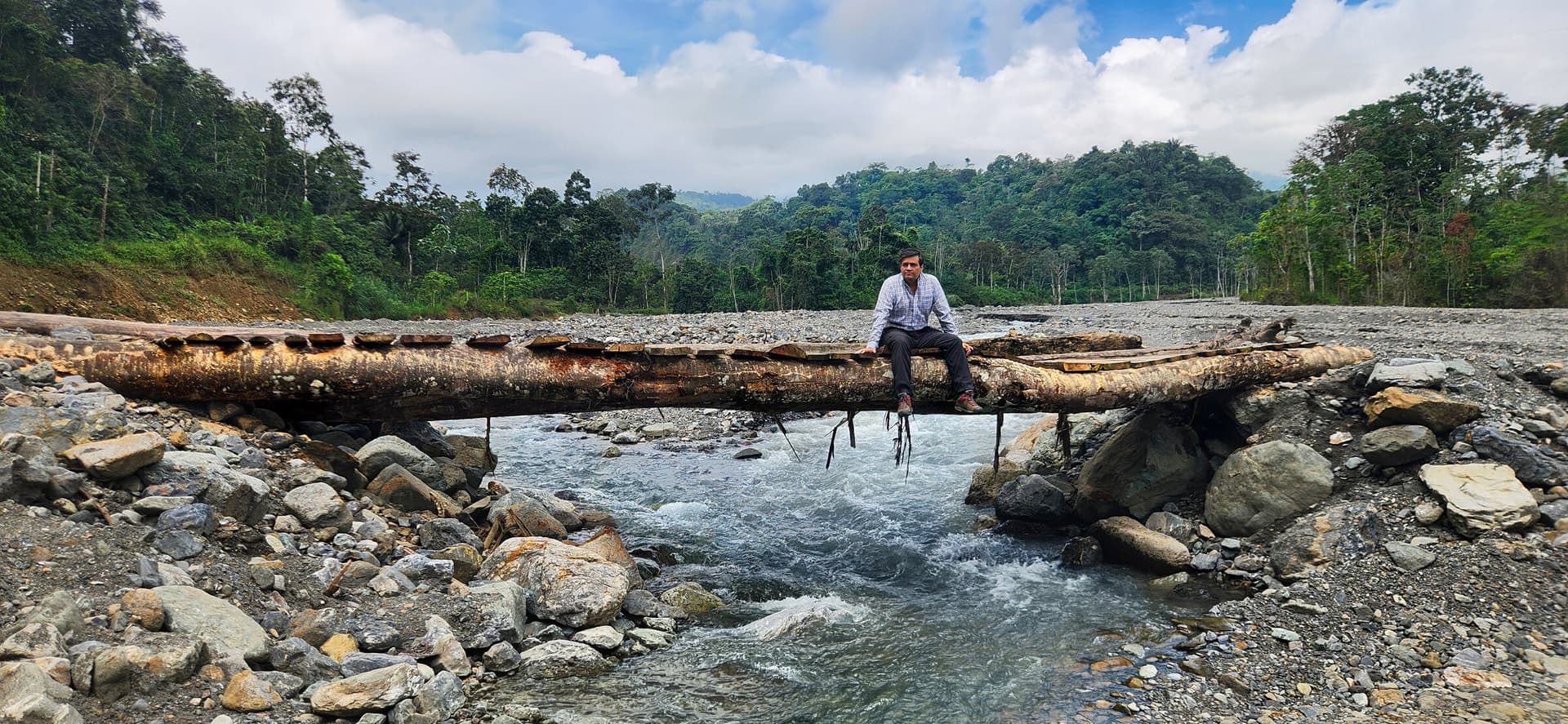 Edgar Landívar sentado sobre un puente de troncos que cruza un río de montaña, rodeado de selva